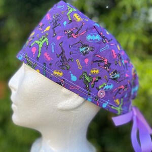 May include: Purple surgical scrub hat with a colorful print of Batman villains. The hat has a button and a purple tie.