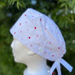 May include: White scrub hat with a pink and red heart pattern. The hat has a button and ties at the back.