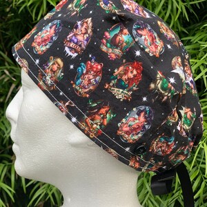 May include: Black scrub hat with a colorful print of various Disney princesses in a whimsical style. The hat has a button on top and a tie at the back.
