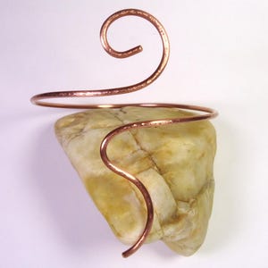 Può includere: Un bracciale in rame con un design a spirale, appoggiato su una pietra di colore chiaro. Il bracciale avvolge la pietra, mostrando la sua texture martellata e la calda tonalità marrone-rossastra. La pietra ha una forma naturale e organica.