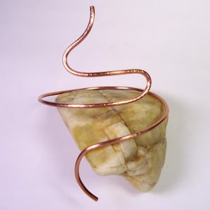 Può includere: Scultura in pietra naturale con una roccia chiara e strutturata con un design in filo di rame. Il filo si avvolge attorno alla pietra e si estende verso l'alto in una forma curva e artistica. La scultura è su sfondo bianco.
