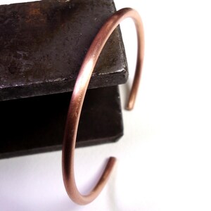 Può includere: Un semplice bracciale a polsino aperto in rame. Il bracciale è una fascia curva solida con estremità aperte. Il rame ha una calda tonalità marrone-rossastra. Il bracciale è mostrato su una superficie scura e strutturata.