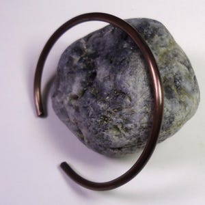 Può includere: Un bracciale a polsino color rame poggia su una roccia screziata grigia e blu su sfondo bianco. Il bracciale ha un design semplice e aperto, con estremità arrotondate. La roccia ha una consistenza ruvida.