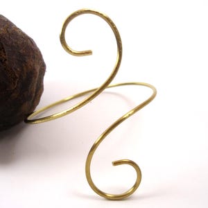 Op de afbeelding: Een gouden metalen armband met een gedraaid ontwerp. De armband heeft twee lussen aan de bovenkant en een enkele lus aan de onderkant.