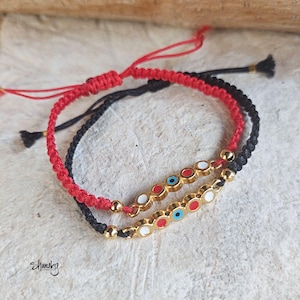 May include: Two handmade bracelets. One is red, the other is black. Both have a woven design. The bracelets feature gold-colored beads and a row of small, colorful eye-shaped charms with red, white, and blue accents. The bracelets are on a light-colored surface.