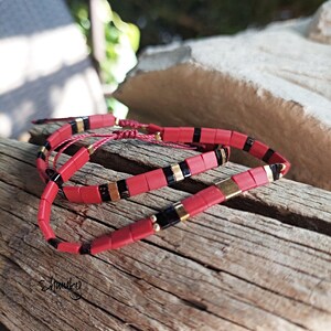 Puede incluir: Una pulsera roja con detalles negros y dorados, colocada sobre un trozo de madera envejecida. La pulsera está hecha de pequeñas cuentas rectangulares. El diseño incluye rayas negras y elementos metálicos dorados. La pulsera está anudada con un cordón rojo.