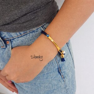 Könnte beinhalten: Goldfarbenes Armband mit blauem Kordel und einem kleinen blauen Augenanhänger. Das Armband hat ein goldenes Stangendesign und eine Quaste mit einer kleinen Perle. Das Wort "Ehmuky" steht auf dem Handgelenk.