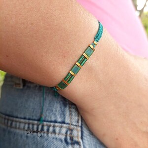 May include: A teal and gold bracelet with rectangular and cylindrical beads. The bracelet is made of teal cord and has gold-coloured metal accents. The bracelet is worn on a wrist, showcasing the design and colour combination. The bracelet is a piece of jewellery.