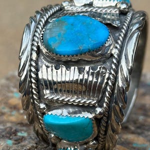 May include: A wide silver ring featuring three bright blue turquoise stones set in serrated bezels. The band is decorated with twisted wire rope detailing, vertical silver bars, and leaf-like engravings along the edges of the metal surface.