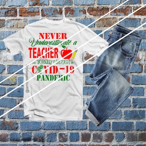 Puede incluir: Camiseta blanca con el texto "Never Underestimate a Teacher who worked through the COVID-19 Pandemic" en letras verdes y rojas. Una manzana roja y un lápiz están incluidos en el diseño. El texto "@iamtshirtplus" está en la esquina inferior izquierda.