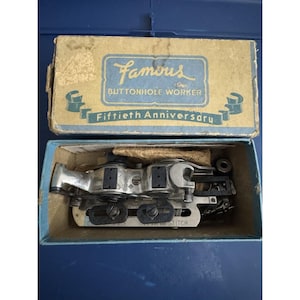 May include: An open box containing a vintage Famous Buttonhole Worker sewing machine attachment. The box is tan and blue with text that reads "Famous Buttonhole Worker" and "Fiftieth Anniversary". The attachment is silver and black.