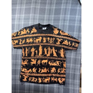 Pode incluir: Camiseta preta com um padrão laranja de inspiração grega. O design inclui figuras, cavalos e elementos arquitetônicos. A camiseta tem mangas curtas e gola redonda. A camiseta está colocada em uma superfície quadriculada.