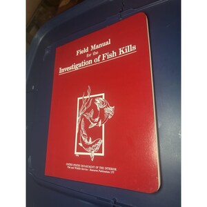 May include: A red field manual titled "Field Manual for the Investigation of Fish Kills." The cover features a white illustration of two fish within a rectangular frame, with text from the United States Department of the Interior.