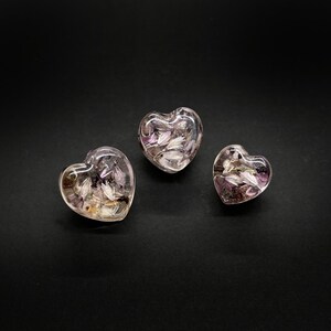 May include: Three heart-shaped resin objects with embedded dried flowers in shades of purple, white, and brown. The clear resin allows the flowers to be visible. The hearts vary slightly in size and are set against a black background.