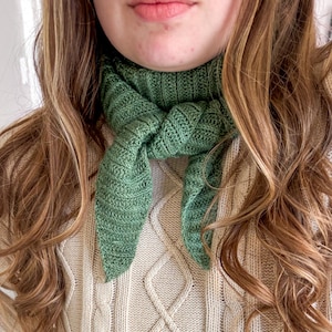 Pode incluir: Um cachecol verde tricotado amarrado ao redor do pescoço. O cachecol é de cor verde lisa e tem uma aparência texturizada. A pessoa está usando um suéter de malha creme e jeans azul.