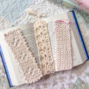 May include: Three handmade crochet bookmarks in cream color, resting on an open book. The bookmarks feature intricate openwork designs and a tassel. One bookmark has a pink ribbon. The book is open to page 220.