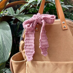 May include: A brown canvas backpack with a pink crocheted bow tied to the handle. The backpack has a large pocket on the front and a zipped pocket on the side.