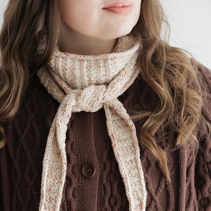 May include: A brown cable-knit cardigan with wooden buttons and a cream-coloured scarf. The scarf is knotted at the neck and has a speckled pattern with hints of red. The cardigan has a textured design.