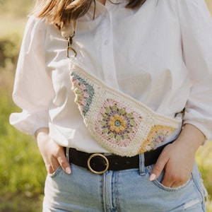 Op de afbeelding: Crèmekleurige gehaakte fanny pack met een bloemenpatroon in pastelkleuren. De tas heeft een lange riem en wordt over het lichaam gedragen. De persoon draagt een wit overhemd, een blauwe spijkerbroek en een zwarte riem met een gouden gesp.