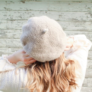 May include: A beige crocheted beret with a pointed top. The beret is worn by a person with long, wavy, light brown hair.