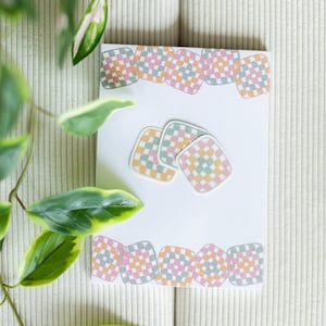 May include: A white card with a border and three square stickers, all featuring a pastel checkered pattern in pink, orange, and green. The card is on a textured, off-white surface, with green and white leaves in the foreground.