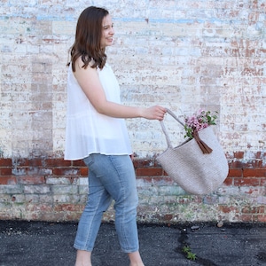 May include: A woman wearing a white sleeveless top, blue jeans, and black sandals is holding a beige crocheted tote bag with a brown tassel. The bag is filled with pink flowers.