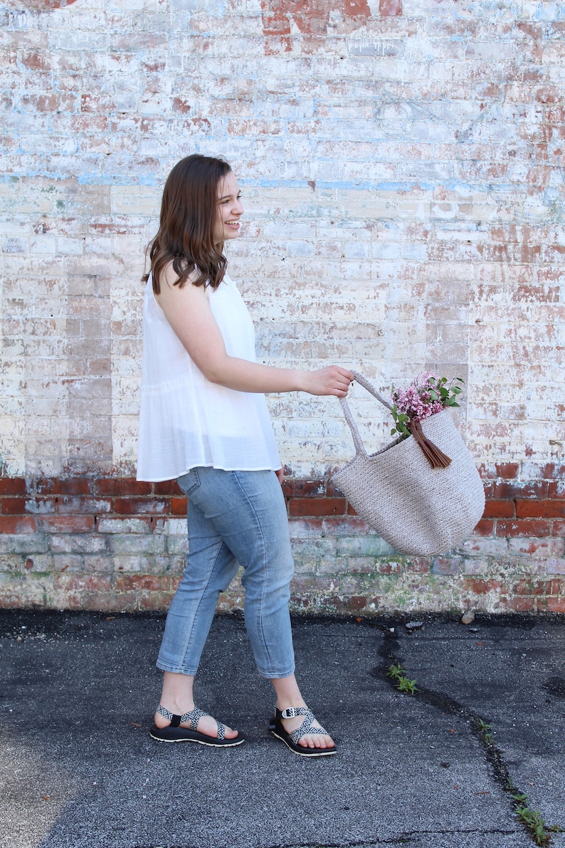 May include: A woman wearing a white sleeveless top, blue jeans, and black sandals is holding a beige crocheted tote bag with a brown tassel. The bag is filled with pink flowers.