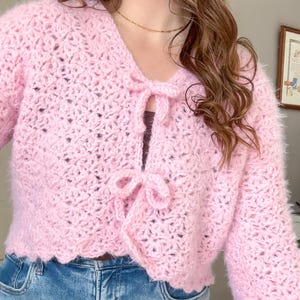 May include: A light pink, open-front, crocheted cardigan with long sleeves. The sweater features a V-neckline and is tied with two bows. The bottom hem has a scalloped edge. The cardigan is made of a fuzzy yarn.