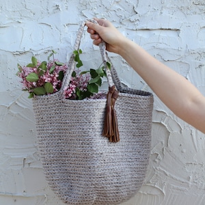 May include: A light gray crocheted tote bag with a brown leather tassel. The bag is filled with a bouquet of pink and white flowers.