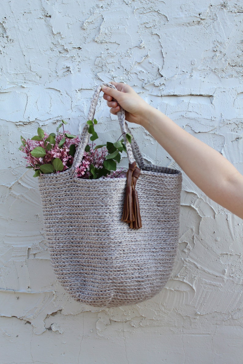 May include: A light gray crocheted tote bag with a brown leather tassel. The bag is filled with a bouquet of pink and white flowers.