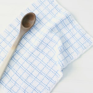 May include: A white crocheted dishcloth with blue stripes. The dishcloth is folded and a wooden spoon is lying on top of it.