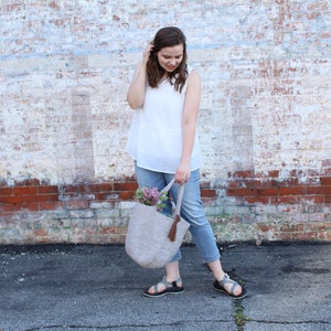 May include: A woman wearing a white sleeveless top and blue jeans is holding a light gray crocheted tote bag with a brown tassel. The bag is filled with purple flowers.