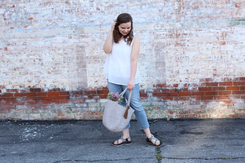 May include: A woman wearing a white sleeveless top and blue jeans is holding a light gray crocheted tote bag with a brown tassel. The bag is filled with purple flowers.