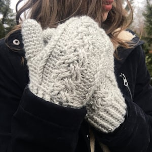 May include: A pair of gray knitted mittens with a cable knit pattern. The mittens are held by a person wearing a black jacket.