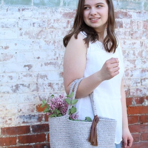 May include: A woman in a white tank top holds a crocheted tote bag with a brown leather tassel. The bag is filled with a bouquet of purple flowers.