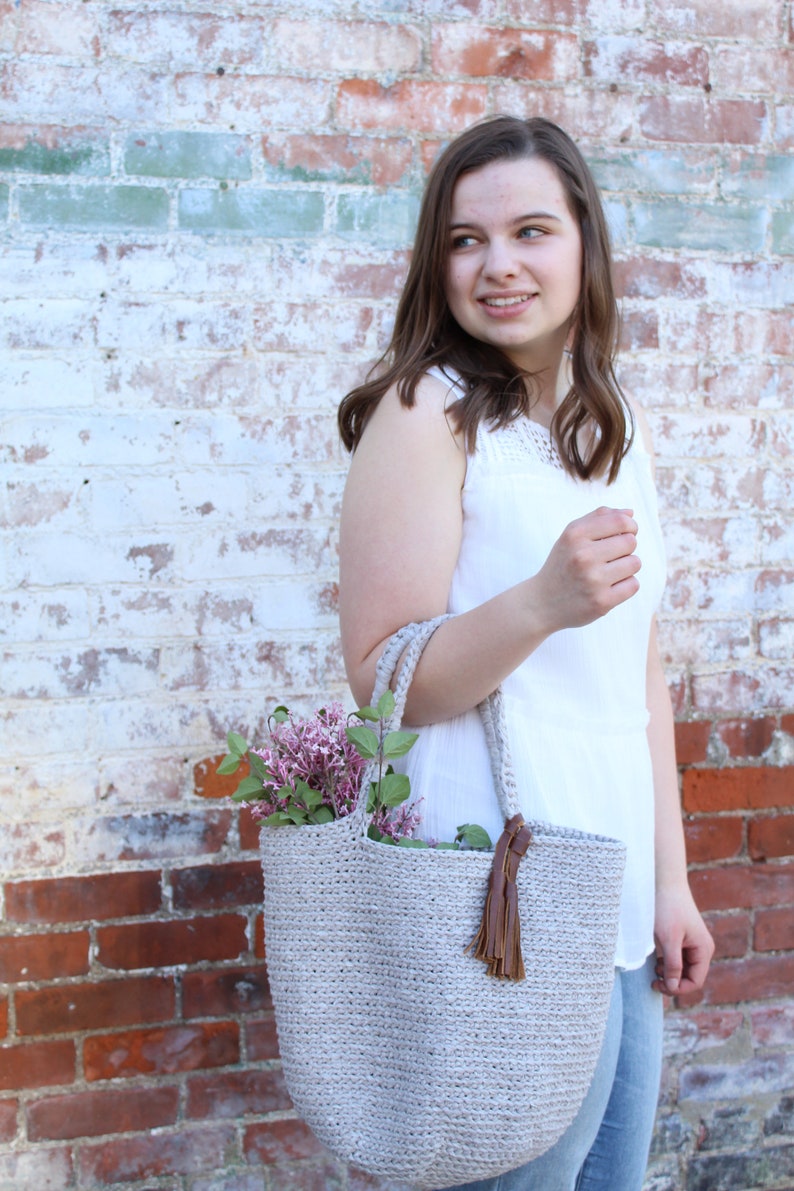 May include: A woman in a white tank top holds a crocheted tote bag with a brown leather tassel. The bag is filled with a bouquet of purple flowers.