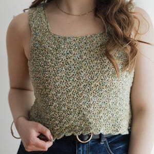 May include: A sleeveless crochet top featuring a square neckline and a textured, multi-colored yarn pattern in shades of sage green, tan, and subtle blue. The garment has a scalloped hemline and is worn with dark blue denim jeans and a black belt.