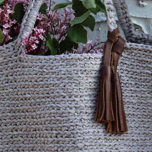 May include: A light gray crocheted tote bag with brown leather handles and a tassel. The bag is filled with a bouquet of purple flowers.