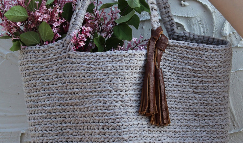 May include: A light gray crocheted tote bag with brown leather handles and a tassel. The bag is filled with a bouquet of purple flowers.