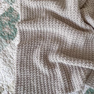 May include: A beige crocheted throw blanket with a textured pattern. The blanket is folded and draped over a patterned surface.