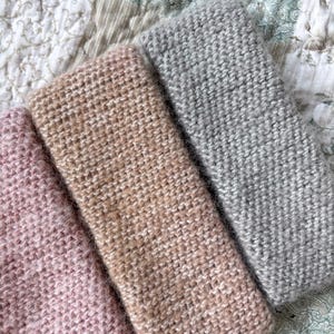 Könnte beinhalten: Drei gestrickte Stirnbänder in Rosa-, Beige- und Grautönen. Die Stirnbänder sind aus weichem, strukturiertem Garn gefertigt und ordentlich gefaltet. Die Stirnbänder werden auf einem Stoffhintergrund mit Muster präsentiert.