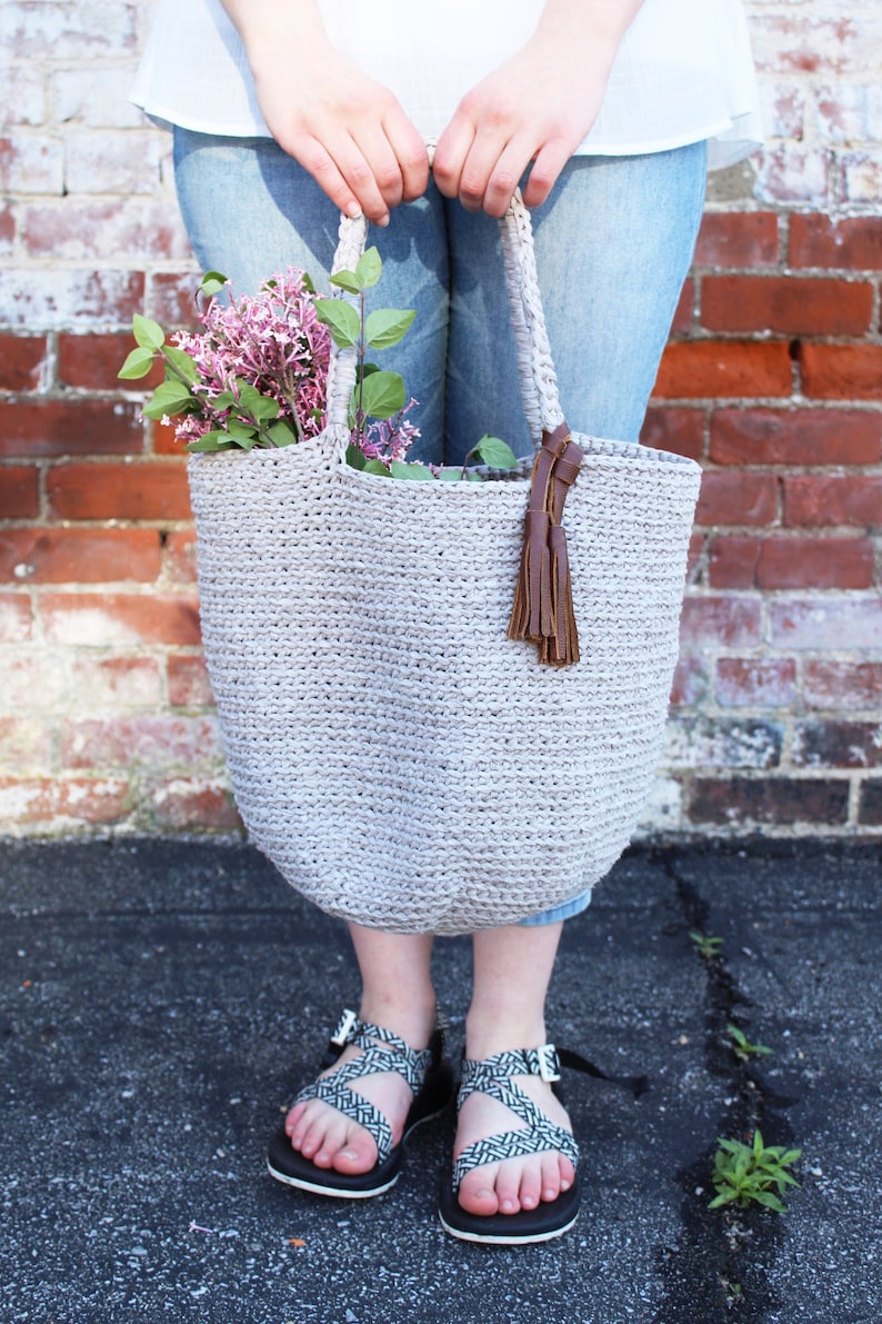 May include: A gray crocheted tote bag with brown leather straps and tassel. The bag is filled with a bouquet of pink flowers.