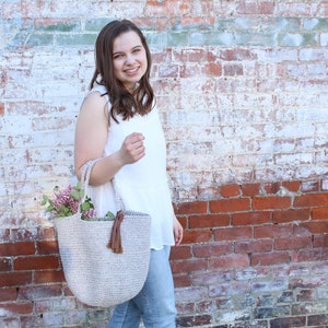 May include: A woman is holding a light beige crocheted tote bag with a brown tassel. The bag is filled with pink flowers. She is standing in front of a white brick wall with red brick accents.
