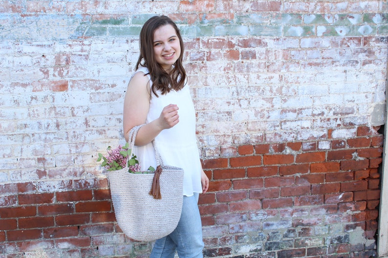 May include: A woman is holding a light beige crocheted tote bag with a brown tassel. The bag is filled with pink flowers. She is standing in front of a white brick wall with red brick accents.