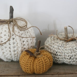 May include: Three crocheted pumpkins in white, gray, and mustard yellow. The pumpkins have twig stems and are tied with twine.