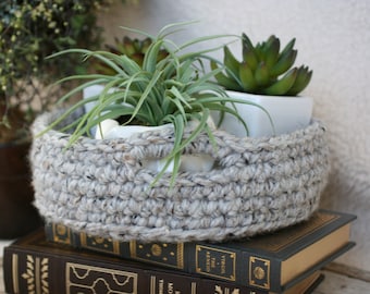 May include: A grey crocheted basket with two potted succulents. The basket has a handle and is sitting on top of a stack of books.