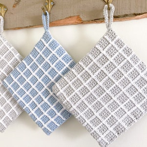 Może przedstawiać: Cztery szydełkowe uchwyty do garnków wiszące na haczykach. Dwa są szare z białymi kwadratami, a dwa są niebieskie z białymi kwadratami.