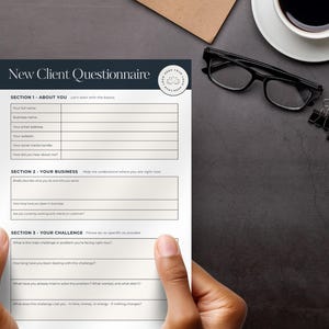 May include: A printed "New Client Questionnaire" form with sections for personal and business information. The form is held by hands and is on a dark surface with a coffee cup, glasses, and a notepad.