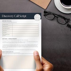May include: A person holding a printed "Discovery Call Script" on a dark surface. The script has a header and sections for information. Other items on the surface include a cup of coffee, glasses, a notepad, and binder clips.