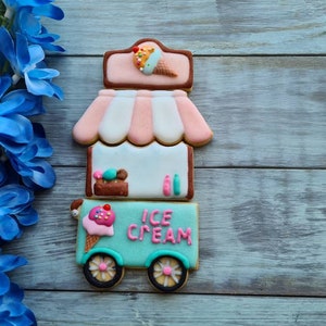 May include: A whimsical ice cream cart cookie with a pink and white striped awning, and the words "ICE CREAM" in pink. The cookie is decorated with an ice cream cone and other ice cream treats. Blue flowers are in the background.